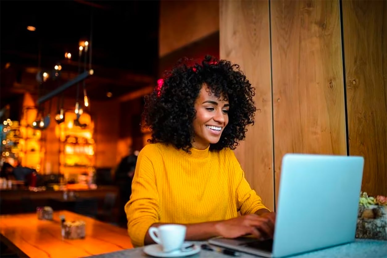 Woman is sitting in a cafe, smiling and typing into her laptop