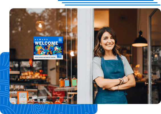 A woman standing in the door of her small business with a POP sticker welcoming American Express in the window.