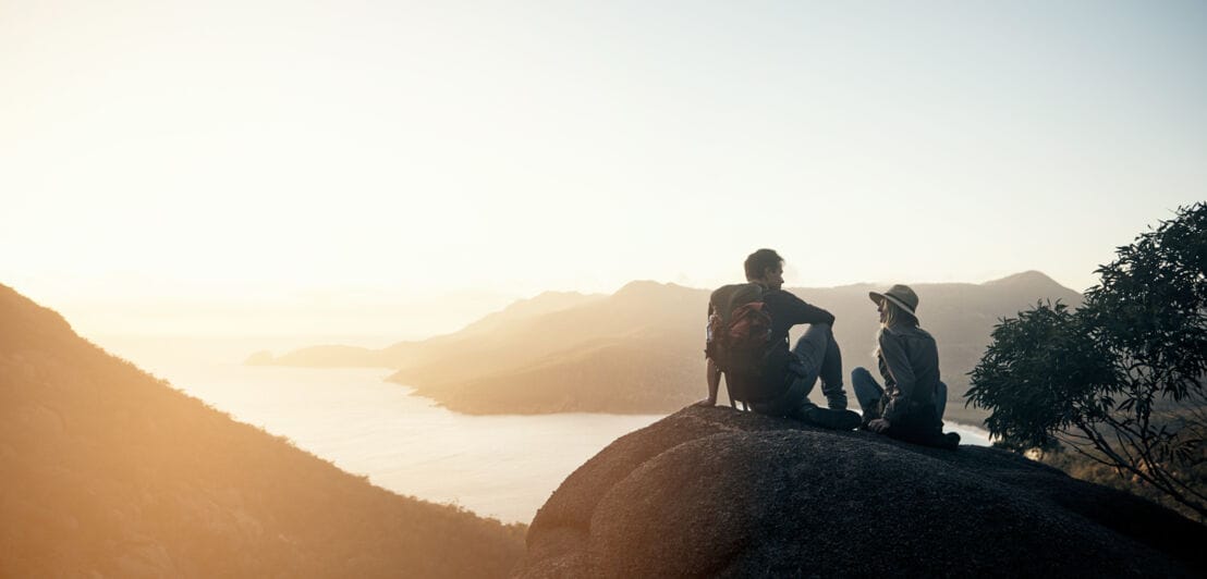Ein Paar sitzt auf einem Felsvorsprung über einer Bucht im Sonnenuntergang Ein Paar sitzt auf einem Felsvorsprung über einer Bucht im Sonnenuntergang