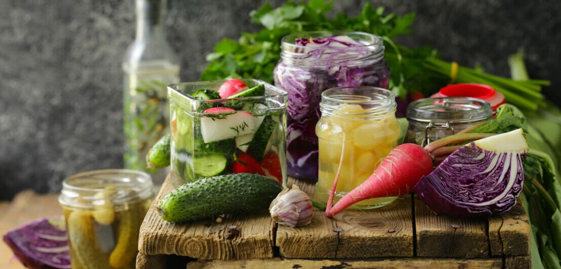 Verschiedene eingelegte Gemüse in Gläsern, darunter Gurken, Rotkohl und Zwiebeln, auf einem rustikalen Holztisch mit frischem Gemüse im Hintergrund. Verschiedene eingelegte Gemüse in Gläsern, darunter Gurken, Rotkohl und Zwiebeln, auf einem rustikalen Holztisch mit frischem Gemüse im Hintergrund.