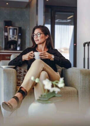 Eine Frau sitzt mit einer Kaffeetasse in einem Sessel in einer belebten Lounge, neben ihr ein Reisekoffer.